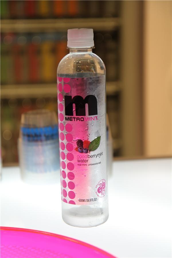 Close-up of a Metromint goodberrymint water bottle with a pink dotted label reading real mint, unsweetened, standing on a white surface with blurred shelves behind it.