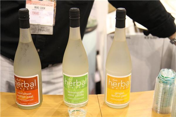 Three bottles of Ayala's Herbal Water in cinnamon orange peel, lemongrass mint vanilla and ginger lemon peel stand on a table in front of Mike Cook, whose badge is visible, with a stack of cups beside them.