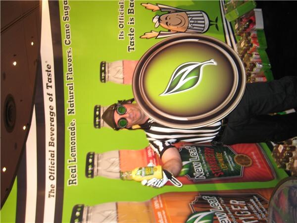 Sideways photo of a person in a referee shirt, goggles and cap holding a bottle of New Leaf tea and a large leaf-logo shield in front of a green New Leaf banner.