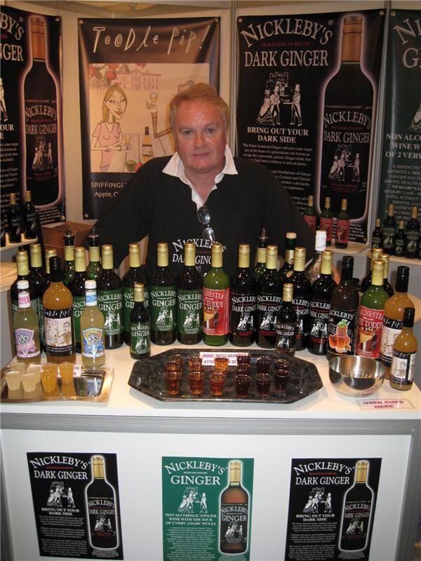 A man stands behind a counter lined with Nickleby's Dark Ginger and Ginger bottles, with sample cups on a tray and Toodle Pip and Nickleby's banners behind him.
