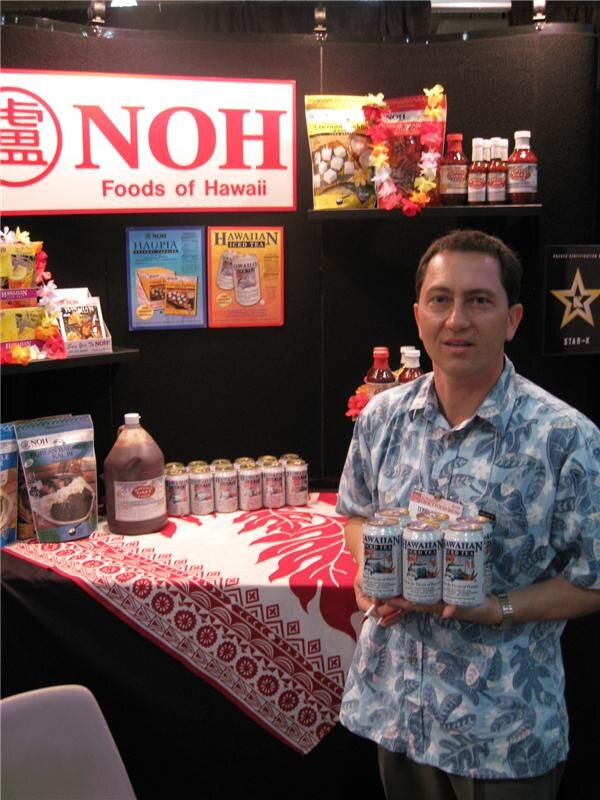 A man in a blue Hawaiian-print shirt holds cans of Hawaiian Iced Tea in front of a NOH Foods of Hawaii sign, with sauces and packaged foods on shelves and a table.
