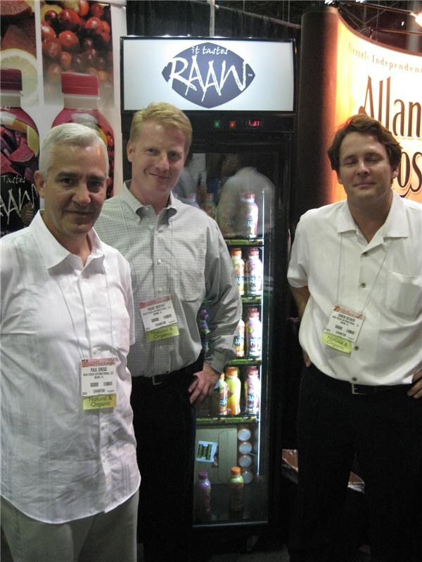 Paul Gregg at left and Trent Moffat at center stand with a third man beside a lit Raaw display cooler filled with bottled drinks. All three wear badges.