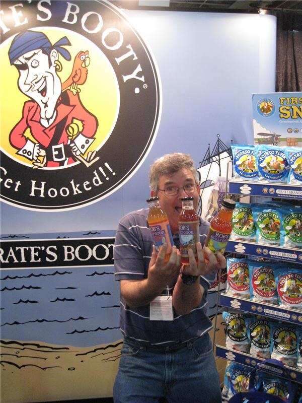 A man with glasses holds three bottled Rob's Really Good drinks in front of a large Pirate's Booty sign. A rack of Potato Flyers chip bags from Pirate Brands stands beside him.