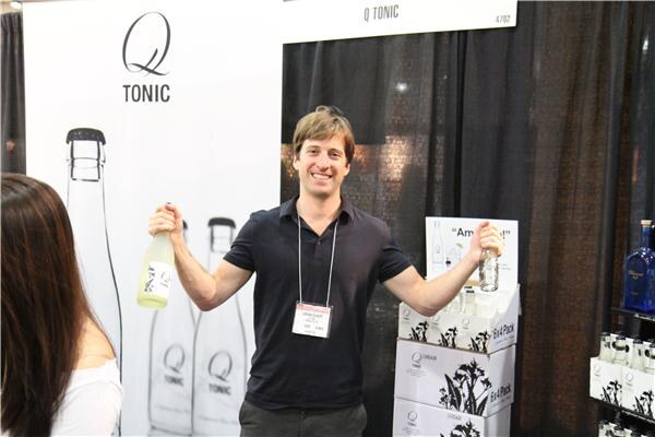 A smiling man wearing a badge holds up two bottles with arms outstretched in the Q Tonic booth, with Q Tonic bottle graphics behind him and cases of Q Tonic stacked beside him.