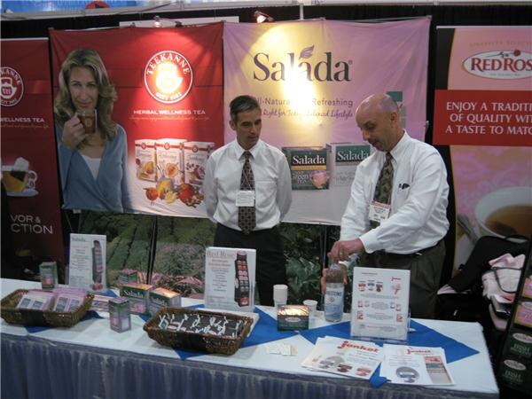 Two men in dress shirts and ties stand behind a table of tea boxes and brochures. Banners for Teekanne, Salada and Red Rose hang behind them.