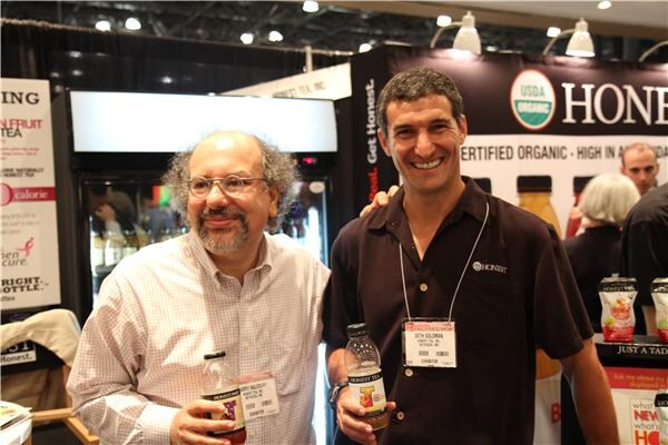 Seth Goldman of Honest Tea, Inc. smiles in a dark shirt and badge beside a bearded man with glasses. Each holds an Honest Tea bottle in front of the Honest Tea booth.