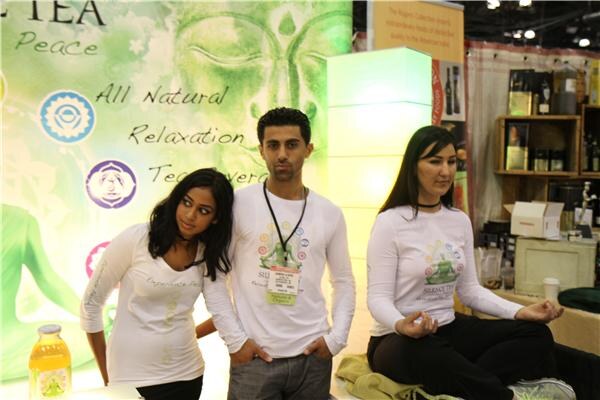 Two women and a man in white Silence Tea shirts stand in front of a green banner reading All Natural Relaxation. One woman sits cross-legged in a meditation pose; a bottle of tea sits at lower left.