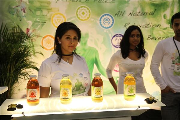 A woman stands behind a lit counter holding four bottles of Silence Tea in orange and yellow. Another woman and a man stand behind her in front of a green All Natural Relaxation banner.