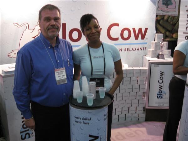 A bearded man in a blue shirt and a woman in a light blue Slow Cow shirt stand together beside a round table with cups and a can. Slow Cow signs and stacked boxes are behind them.