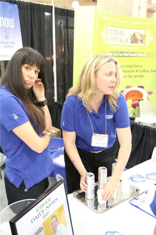 Two women in blue Snow polo shirts stand at a table with cans and cups on a tray. A Snow banner is at upper left and an Ethnic Cottage sign is behind them.