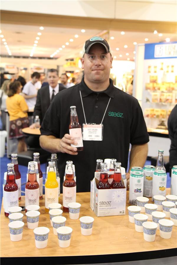 A man in a black Steaz polo and cap holds a bottle of Steaz. In front of him are more Steaz bottles, cans, a four-pack and rows of paper cups on a wooden counter.