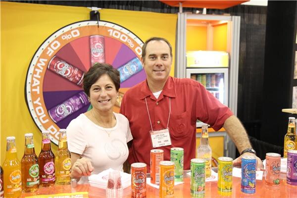 A smiling woman in a white top and a man in a red shirt with a badge stand at a counter of Switch sparkling juice cans and bottles, in front of a spinning prize wheel showing Switch cans.