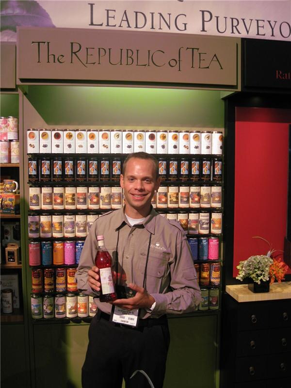A smiling man in a khaki shirt holds a bottle of red tea drink in front of shelves of The Republic of Tea tins under the brand's sign.