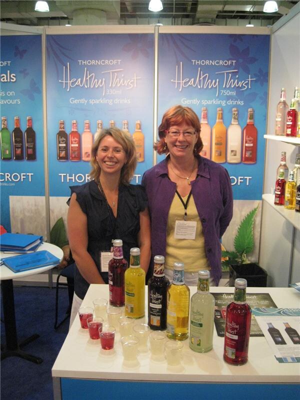 Two smiling women stand behind a table of Healthy Thirst bottles and small sample cups, in front of Thorncroft Healthy Thirst banners showing gently sparkling drinks.