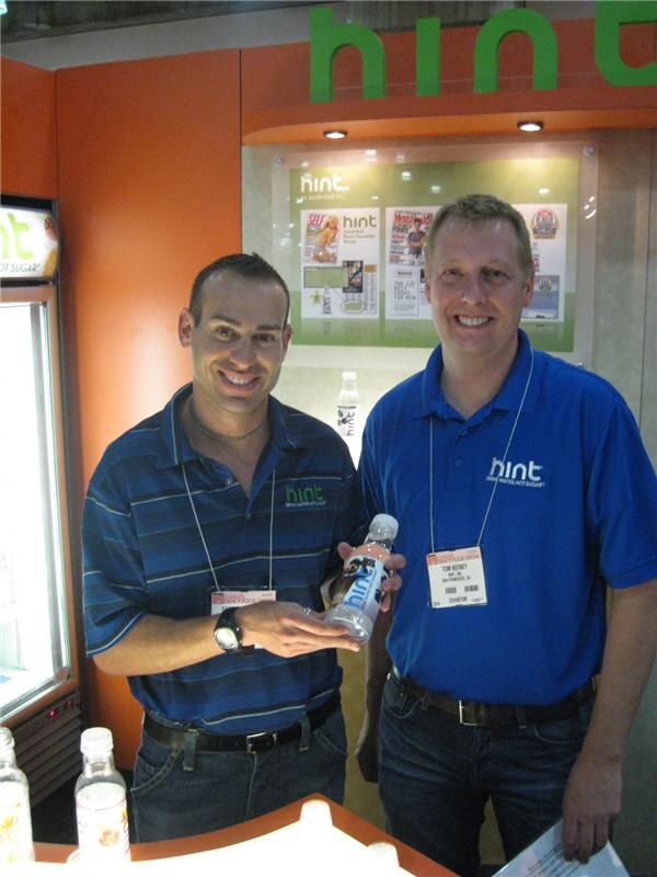 Two smiling men stand at the Hint booth. The man on the right is Tom Keeney, in a blue Hint polo with a badge. The man on the left holds a bottle of Hint water.