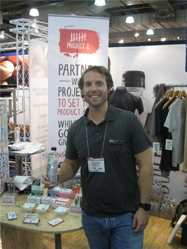 Tyler Merrick of Project 7 smiles and holds a water bottle at the Project 7 booth, with a banner behind him and packaged products and apparel on display.