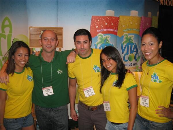 Five people, four in yellow Vita Coco shirts and one man in a green shirt, pose together with badges in front of large Vita Coco coconut water cartons.