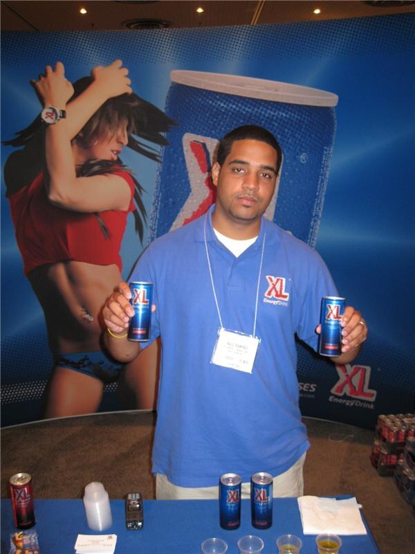 A man in a blue XL polo holds up two cans of XL Energy Drink in front of a blue XL backdrop with a large can graphic, with more cans and sample cups on the table.