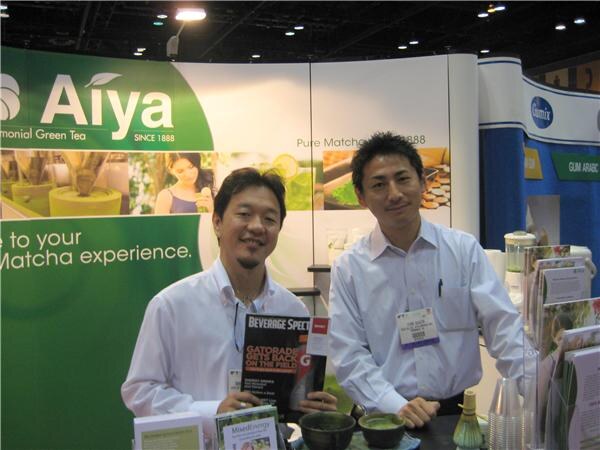 Two smiling men in white shirts stand at the Aiya matcha booth, one holding a Beverage Spectrum magazine, with bowls of green matcha and brochures on the table.