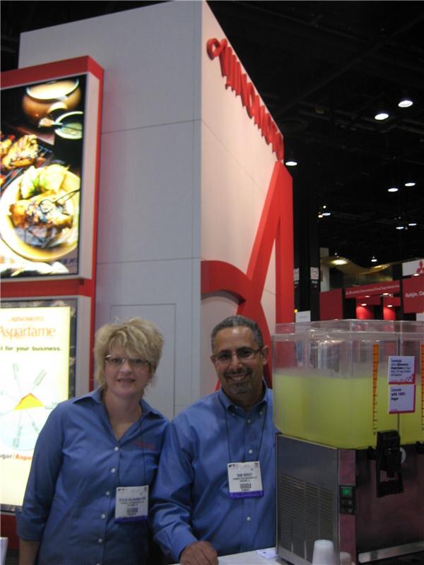 A woman and a man in blue shirts with badges stand beside a beverage dispenser of yellow-green lemonade at the Ajinomoto booth, with food and aspartame displays behind.