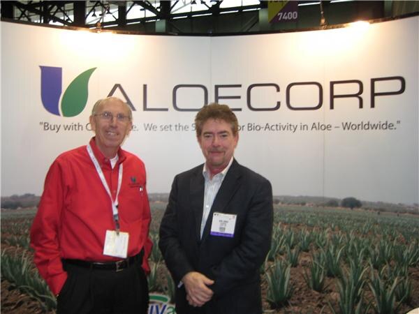 Two men stand side by side in front of an Aloecorp wall graphic showing a field of aloe plants, one in a red shirt and one in a dark blazer, both wearing badges.