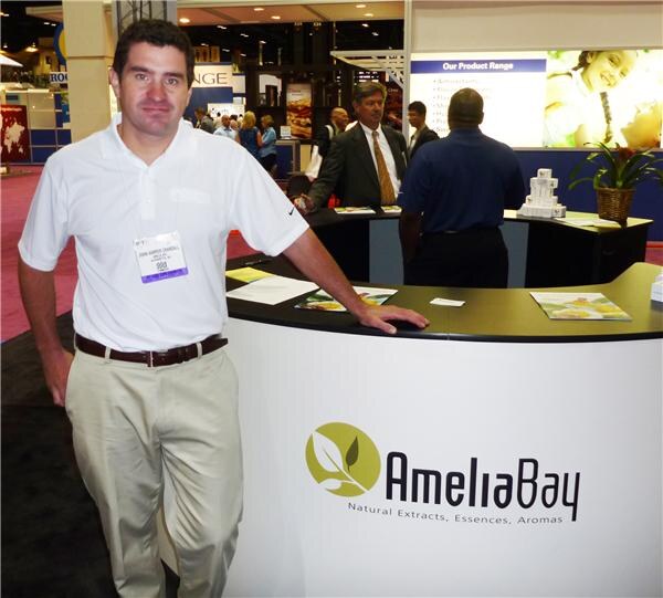 A smiling man in a white polo and khaki pants with a show badge stands beside a curved booth counter with the Amelia Bay logo. Brochures sit on the counter and other attendees stand behind.