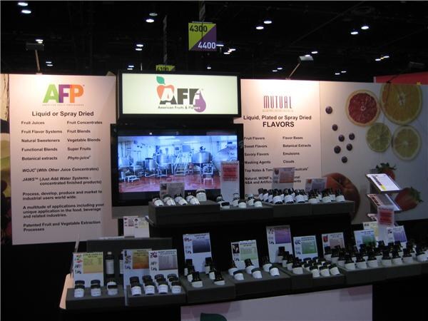 Trade show booth with AFP and American Fruits & Flavors signs, a Mutual flavors panel, a central video screen and rows of small sample bottles on display shelves, with citrus and berry graphics.