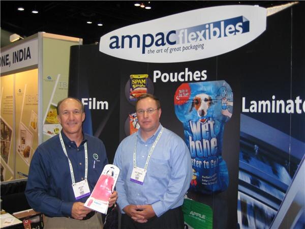 Two men in button-up shirts and show badges stand in front of the Ampac Flexibles booth. One holds a pink pouch. Display graphics show pouches, including SPAM and Wet Bone packages.