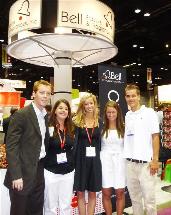 Five smiling people, a man and four women, pose together under a large round Bell Flavors & Fragrances sign. A black Bell banner stands behind them and most wear show badges.