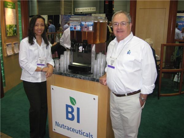 A woman and a man in white shirts with show badges stand beside a wooden counter with a BI Nutraceuticals sign. On the counter are a beverage dispenser and stacks of clear cups.