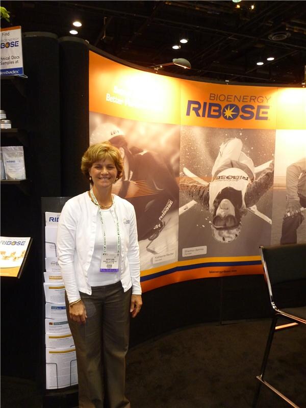 A smiling woman in a white cardigan with a show badge stands in front of a curved orange Bioenergy Ribose display with athlete photos. Brochures and product bags are on shelves beside her.