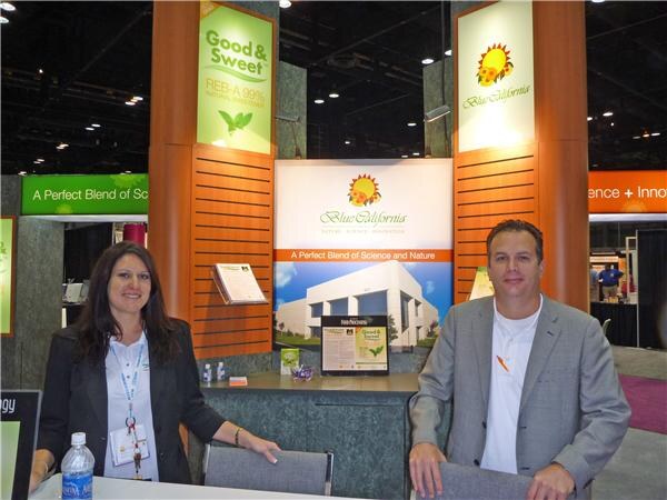A woman in a dark blazer and a man in a gray jacket stand at the Blue California booth. Signs show the Blue California logo and Good & Sweet, with a water bottle on the counter.