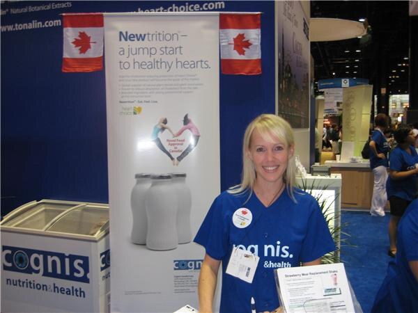 A smiling blonde woman in a blue Cognis shirt stands at the Cognis Nutrition & Health booth. Behind her is a Newtrition banner with Canadian flags and a freezer marked cognis nutrition & health.