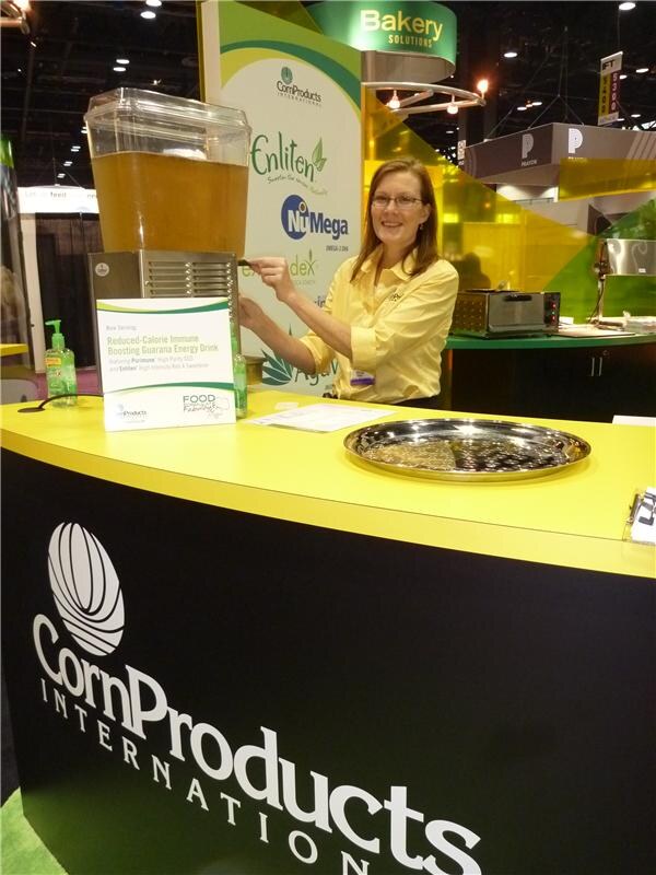 A smiling woman in a yellow shirt and glasses dispenses an amber drink from a dispenser at the Corn Products International booth, with Enliten and NiMega signs behind her.