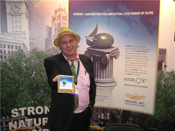 A man in a yellow hat and dark jacket holds a small package toward the camera in front of a Hidrox poster from Creagri, Inc. showing an olive on a column.