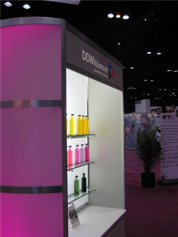 A DDWilliamson booth display with a pink panel and lit shelves holding yellow, pink and green bottles; an IFT 10 sign and other booths are in the background.