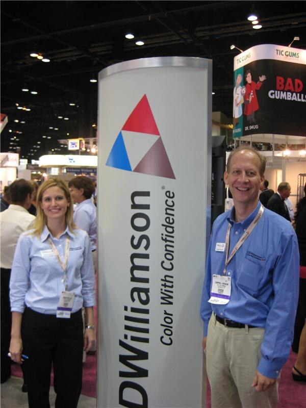 A woman and a man in light blue shirts and badges stand beside a DDWilliamson sign. A TIC Gums booth is visible in the background.