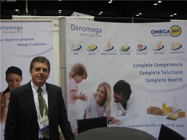 A man in a dark suit and tie with a badge stands in front of Denomega Nutritional Oils and Omega 360 banners showing a family and food icons.