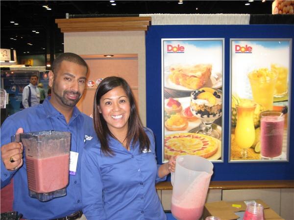 A smiling man and woman in blue shirts hold blenders and pitchers of pink smoothie in front of Dole posters showing desserts and fruit drinks.