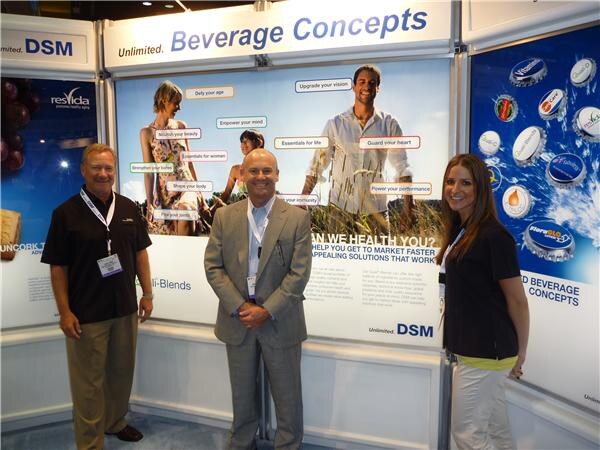 Two men and a woman stand together at the DSM Beverage Concepts booth, with a Resvida sign and a large graphic of people and bottle caps behind them.