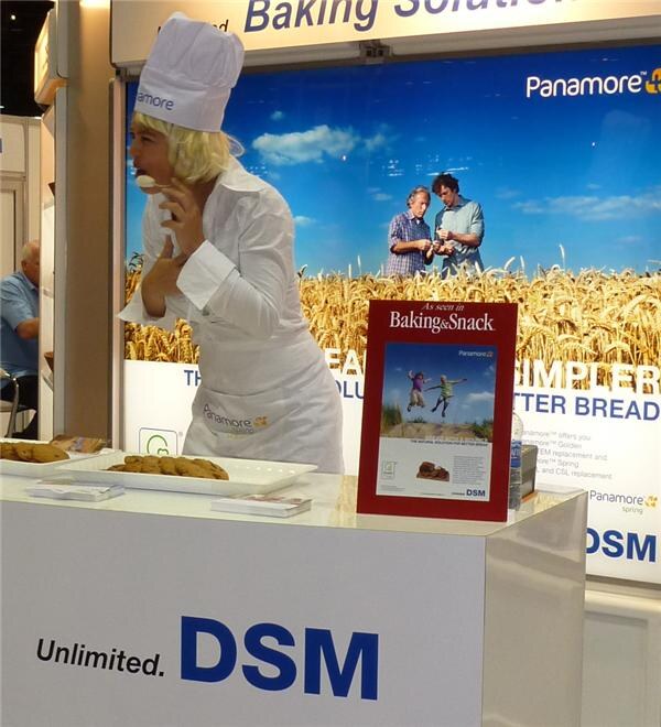 A person in a chef hat, white apron and blonde wig stands at a DSM counter with trays of cookies and a Panamore banner showing a wheat field behind.
