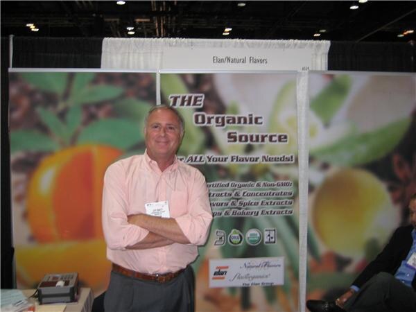 A smiling man in a pink shirt with arms crossed stands in front of an Elan/Natural Flavors banner reading The Organic Source, with citrus images.