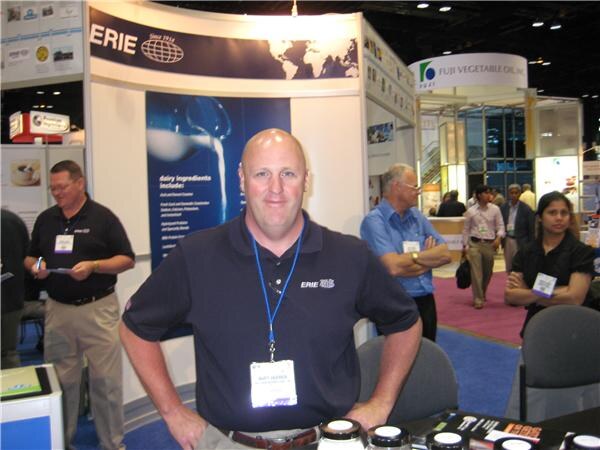 A bald man in a dark ERIE polo with a lanyard and badge stands in front of the ERIE booth. Other people stand behind him, and a Fuji Vegetable Oil sign hangs in the background.