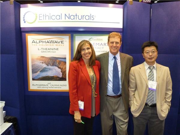 A woman in a red jacket and two men stand together in front of the Ethical Naturals booth, with AlphaWave and PureValue signs on the wall behind them.