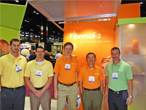 Five men in yellow, orange and green polo shirts with badges stand in a row in front of an orange Fibersol-2 booth wall reading "it's the fiber you want".