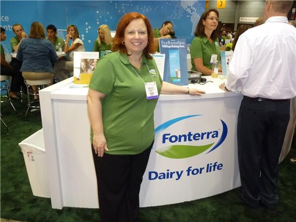 A red-haired woman in a green polo and badge stands beside a white Fonterra counter reading "Dairy for life". Other people in green sit and stand behind it.