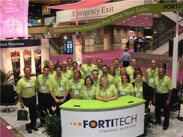 A large group of people in light green polo shirts pose around a curved Fortitech Strategic Nutrition counter. A green banner above reads "Custom Nutrient Premixes that Drive Your Sales".