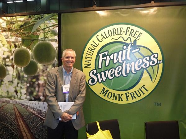A smiling gray-haired man in a tan blazer holds papers in front of a green Fruit-Sweetness Monk Fruit banner. A photo of monk fruit growing hangs on the left.