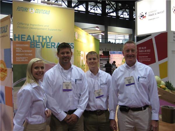 A woman and three men in white shirts with badges stand together in front of a FutureCeuticals sign about healthy beverages. A Germany Trade & Invest sign is on the right.