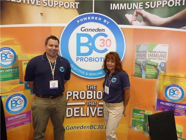 A man and a woman in navy polo shirts with badges stand in front of an orange Ganeden BC30 Probiotic display showing product packages.